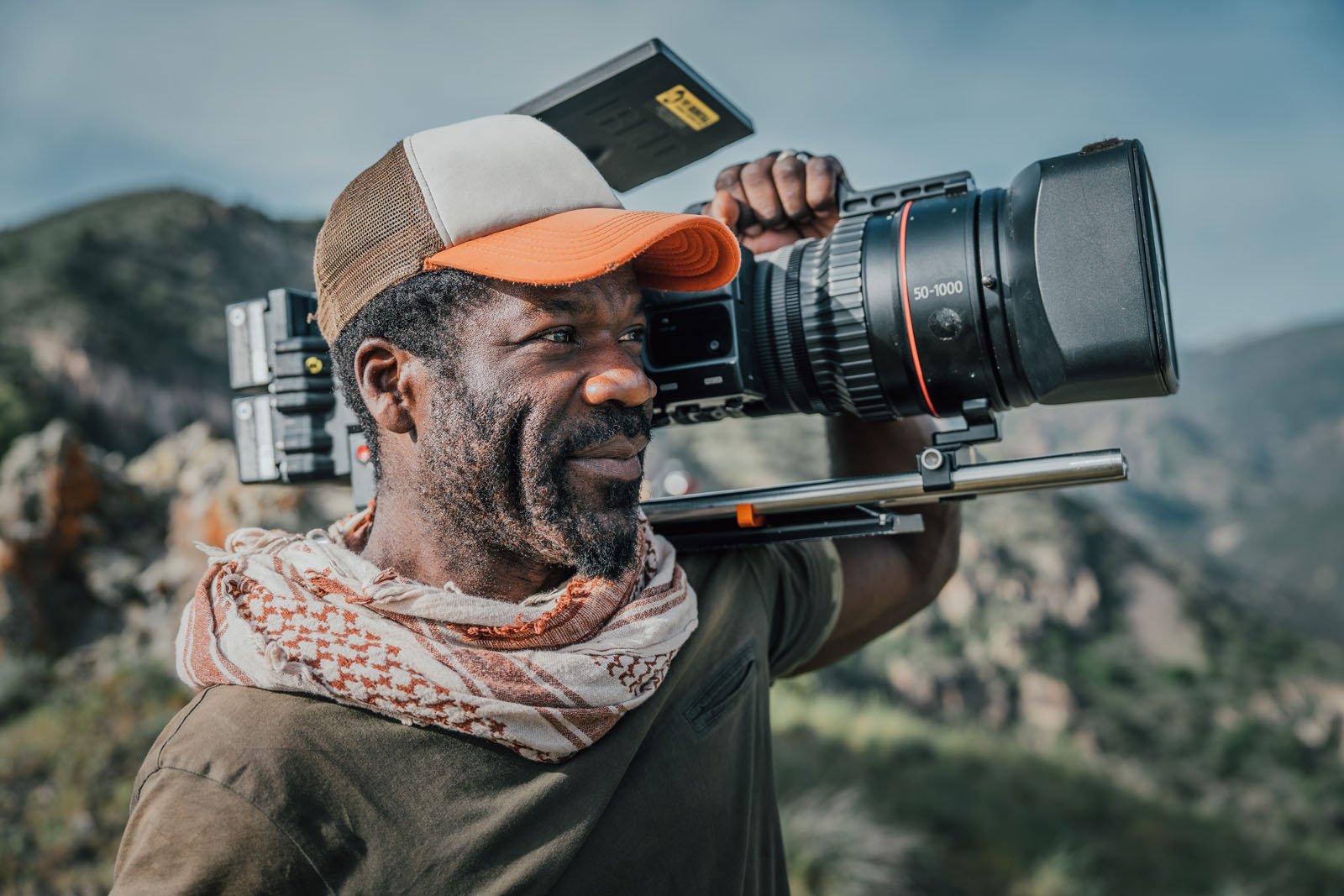 A man wearing a cap and scarf stands outdoors in a mountainous area, smiling and holding a large professional video camera on his shoulder.