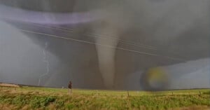 A person stands in a grassy field photographing a large tornado under a dark, cloudy sky. Lightning strikes in the background, and power lines stretch across the scene.