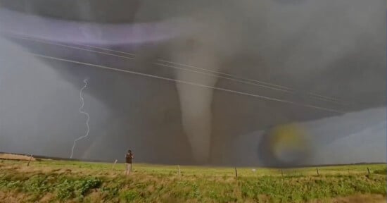 A person stands in a grassy field photographing a large tornado under a dark, cloudy sky. Lightning strikes in the background, and power lines stretch across the scene.