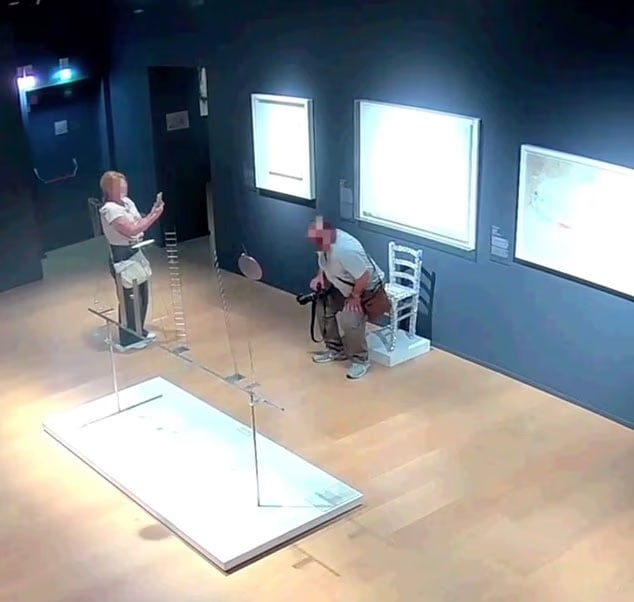 A woman takes a photo of a man sitting on a white chair in an art gallery. The man appears to be posing, and artworks are displayed on the walls behind them. The gallery has dim lighting and a light wooden floor.