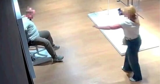 A security camera image shows a person sitting on a chair while another person stands nearby pointing a phone at them. Both faces are blurred, and the scene takes place in a well-lit indoor space with wooden floors.