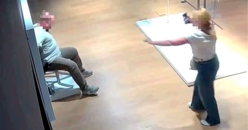 A security camera image shows a person sitting on a chair while another person stands nearby pointing a phone at them. Both faces are blurred, and the scene takes place in a well-lit indoor space with wooden floors.