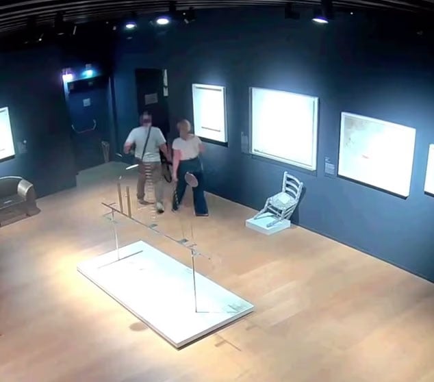 Two people walk through a dimly lit art gallery with wooden floors and framed artworks on dark walls; a chair is displayed on a white platform nearby.