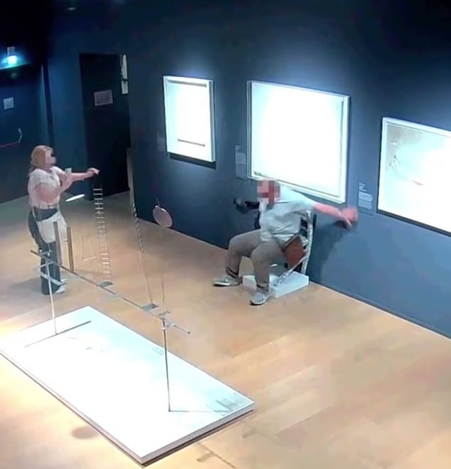 A person sits on a chair attached to a museum wall that is collapsing, while another person stands nearby with arms raised in surprise. Artworks and exhibits are visible in the gallery room.