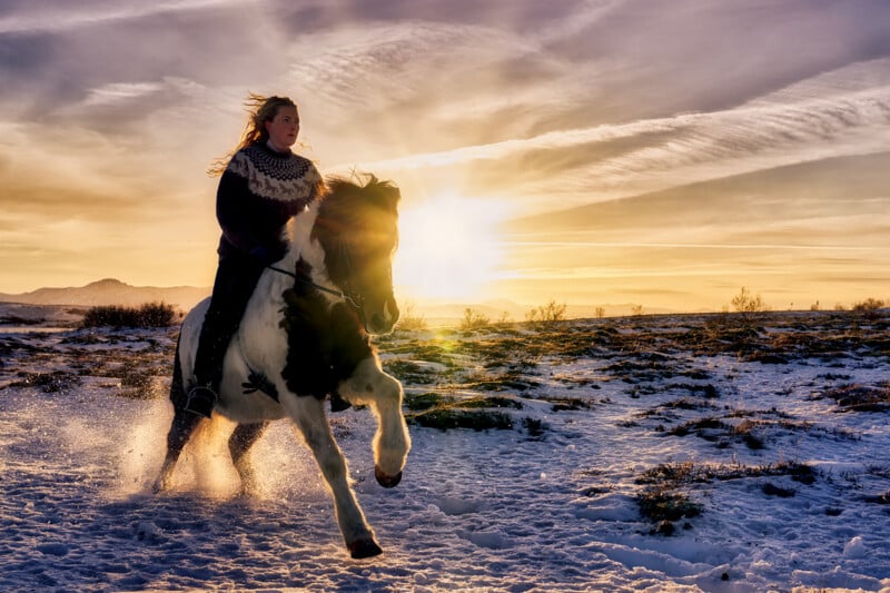 A woman rides a black and white horse across a snowy landscape at sunset, with sunlight streaming behind her and clouds glowing in the sky.