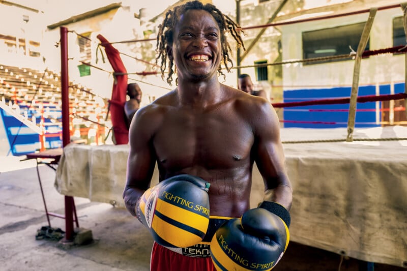 A smiling male boxer with dreadlocks stands in front of a boxing ring outdoors, wearing yellow and black gloves and red shorts. The background shows another person and an empty seating area.