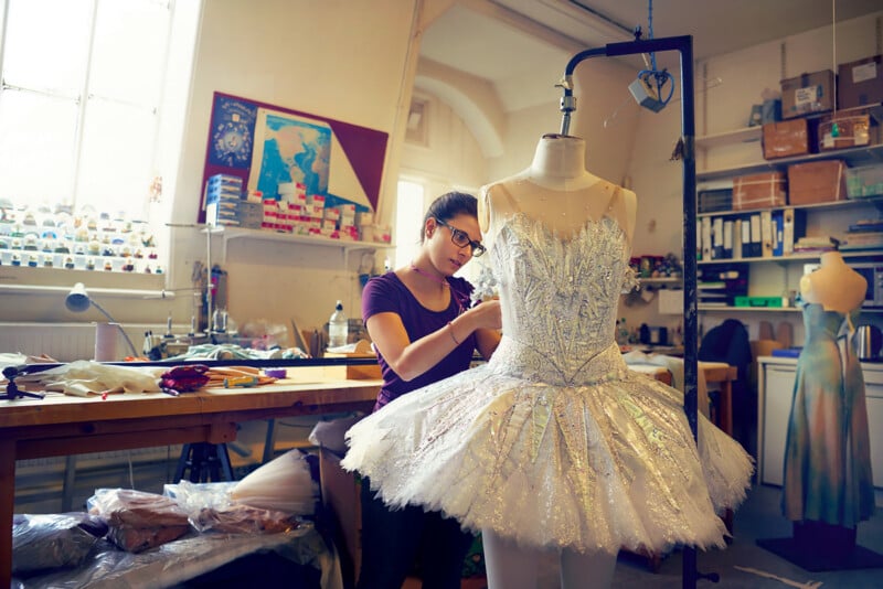 A woman works on a sparkling white ballet tutu displayed on a dress form in a well-lit studio filled with shelves, fabrics, and art supplies.