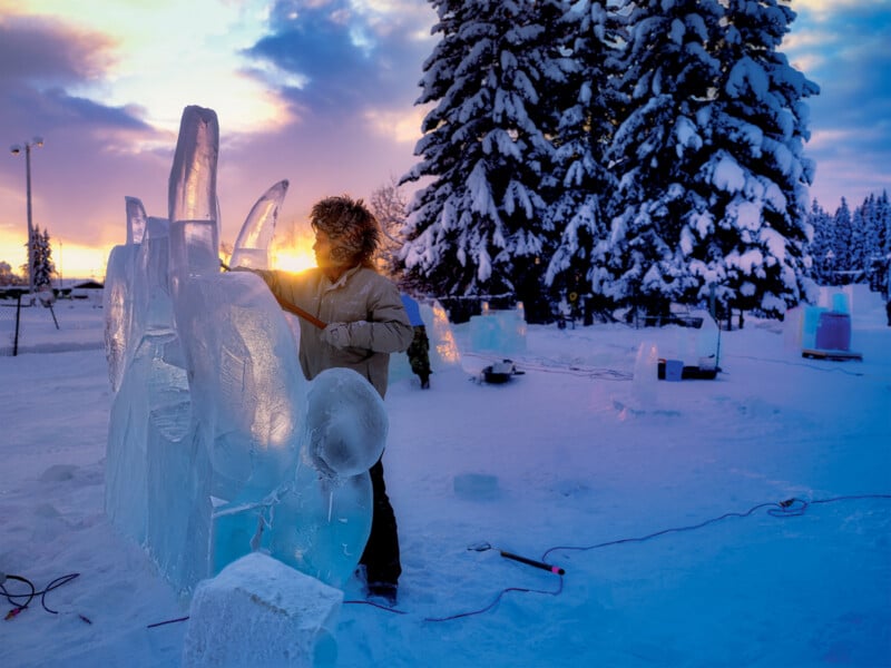 A person in a winter coat carves an ice sculpture outdoors at sunset, surrounded by snow-covered trees and other ice blocks, with tools and equipment scattered on the snowy ground.