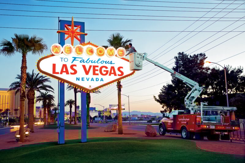 A worker in a cherry picker truck maintains the illuminated "Welcome to Fabulous Las Vegas, Nevada" sign at dusk, with palm trees and a clear sky in the background.