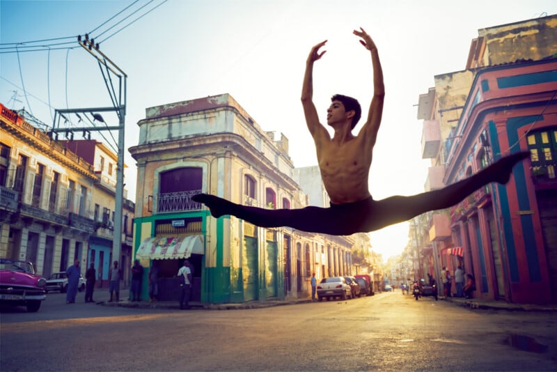 A male ballet dancer performs a high split jump in the middle of a colorful, sunlit street lined with old buildings, as people watch and cars are parked along the sides.