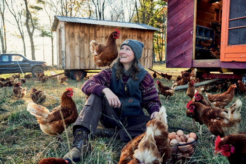 A woman sits on grass surrounded by chickens, smiling and looking up. She wears a knit hat, sweater, and boots. A basket of eggs is by her side, and wooden coops are in the background.
