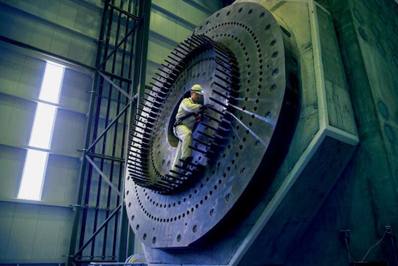 A worker in safety gear uses a tool while standing on a large, circular industrial machine part inside a spacious, modern facility with high ceilings and metal beams.