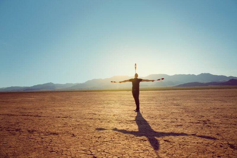 A person is juggling three clubs in an arid, cracked desert landscape at sunset, casting a long shadow with mountains in the background.