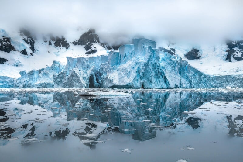 A massive blue glacier with jagged ice rises from calm, reflective water, surrounded by snow-covered mountains and low-hanging clouds in a cold, remote landscape.