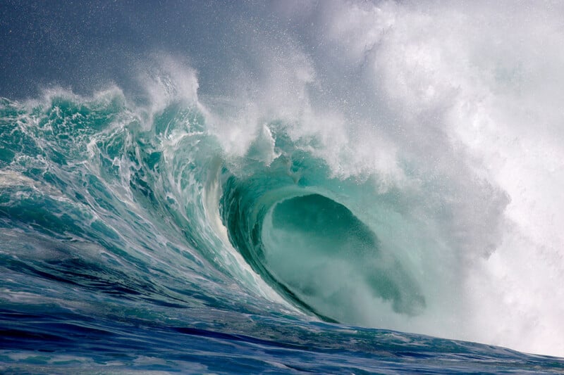 A powerful ocean wave curls, creating a hollow barrel with white spray and foam at the crest, against a backdrop of churning blue water and mist.