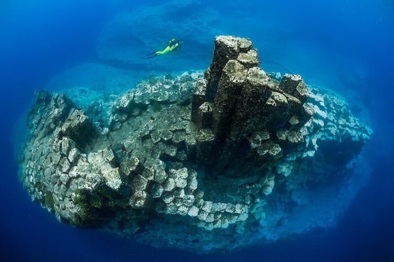 A diver swims above large underwater basalt columns, which rise from the sea floor in geometric, hexagonal shapes, surrounded by clear blue water.