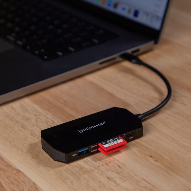 A ProMaster USB hub connected to a laptop on a wooden surface, with an SD card inserted into the hub and a red microSD card partially visible.