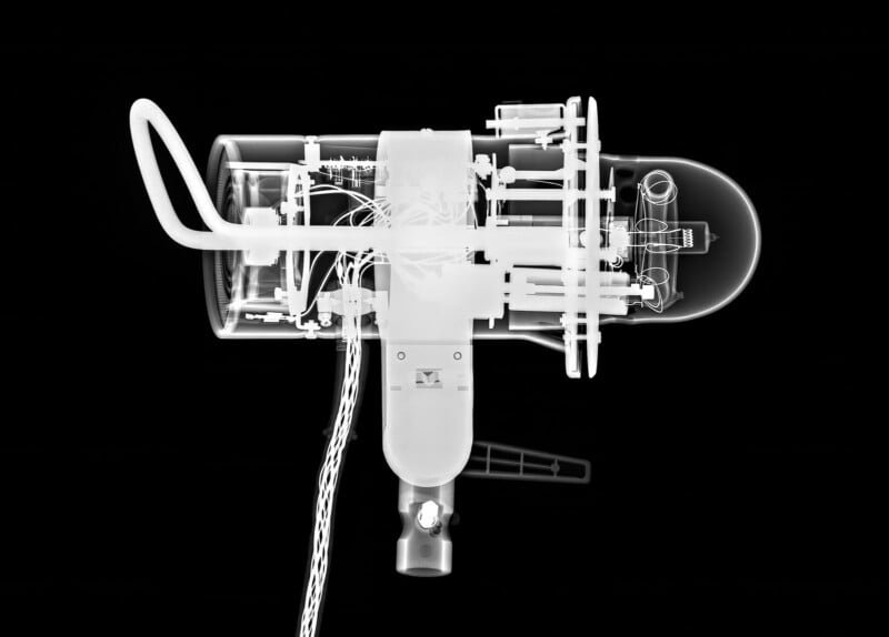 X-ray image of a complex mechanical device with wires, tubes, and various cylindrical and rectangular components, all set against a black background. The internal structures and details are clearly visible.