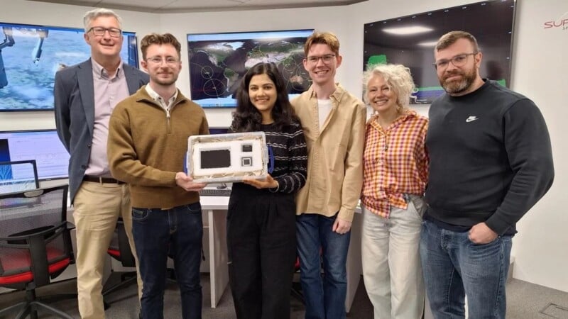 Six people stand together in an office with computer screens behind them; the person in the center holds a white electronic device. The group is smiling and appears to be celebrating an achievement.