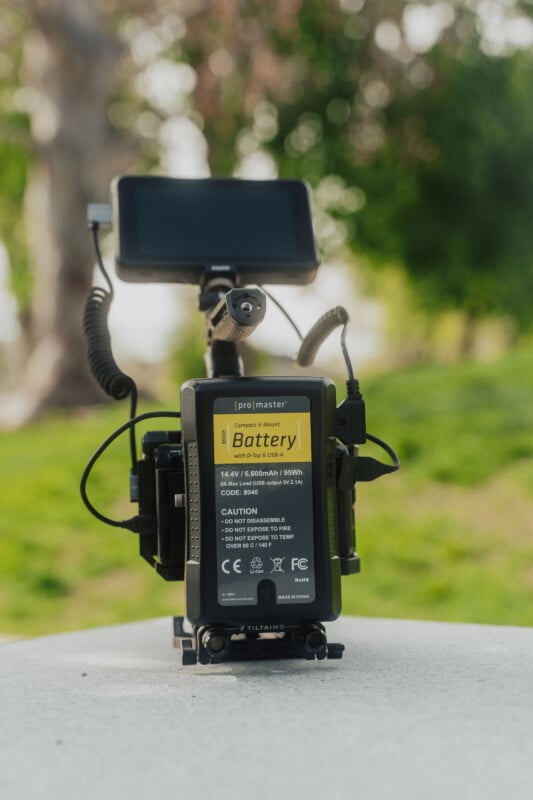 A camera monitor setup with a large ProMaster battery attached, sitting on a white surface outdoors with blurred green grass and trees in the background.