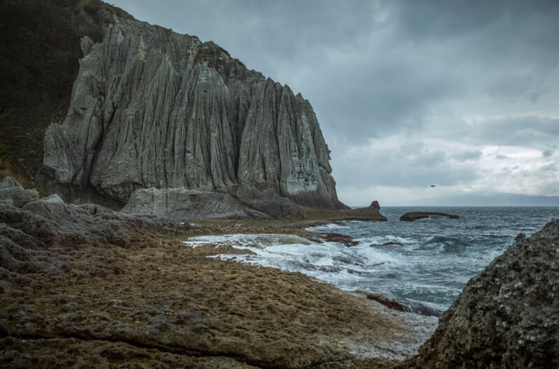 Jagged cliffs with dramatic vertical striations rise beside a rocky shoreline, where waves crash under a cloudy, overcast sky, creating a moody coastal landscape.