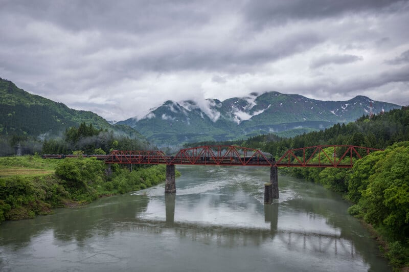 A red steel bridge spans a wide, calm river surrounded by green trees and lush mountains, with mist and clouds hanging over the landscape. A black train crosses the bridge.