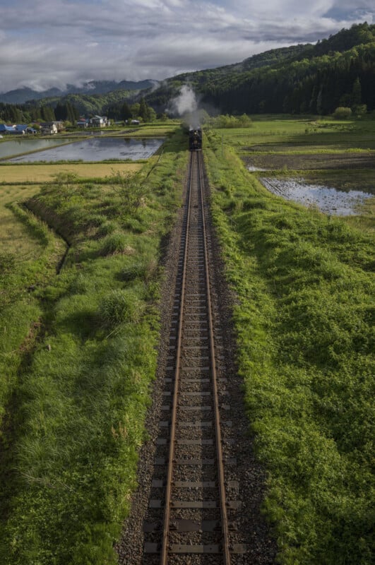 A single train travels along a railway track through lush green fields, with smoke trailing behind. Mountains and a cloudy sky are visible in the background, and water-filled paddies border the track.