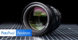 A camera lens rests on a reflective, grid-patterned surface with the "PetaPixel Reviews" logo in the lower left corner. The lens is angled to show the front glass and detailed markings.
