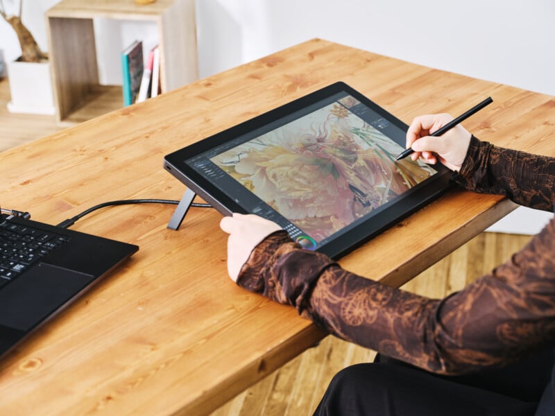 A person uses a stylus to draw a detailed flower on a digital tablet connected to a laptop, seated at a wooden desk in a bright, modern workspace.