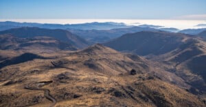 A winding road cuts through dry, rugged mountains under a clear blue sky, with layers of distant hills and mist visible on the horizon.