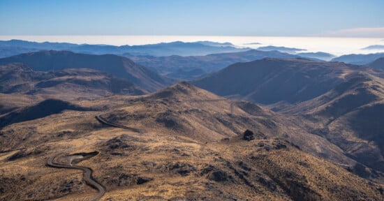 A winding road cuts through dry, rugged mountains under a clear blue sky, with layers of distant hills and mist visible on the horizon.