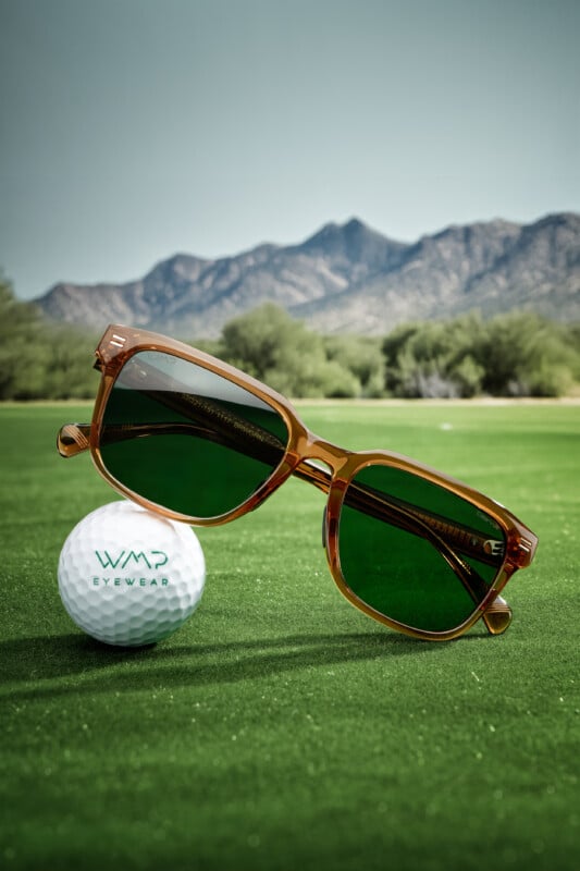A pair of brown-framed sunglasses with green lenses rests on grass next to a golf ball labeled "WMP Eyewear," with mountains and trees in the background under a clear sky.