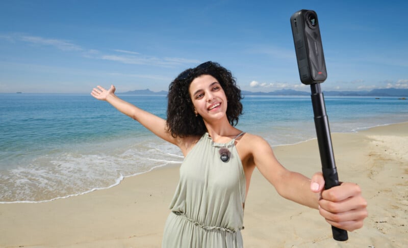 A woman in a light dress stands on a sandy beach, smiling and holding a 360-degree camera on a selfie stick, with one arm outstretched. The ocean and distant mountains are visible under a clear blue sky.