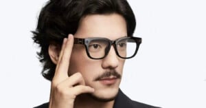 A man with black hair and a mustache is wearing smart glasses and touching the side of the frame with his fingers, as if using a touch control. The background is plain and light-colored.