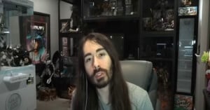 A person with long brown hair and a beard sits in a chair, speaking, with shelves filled with collectibles and action figures in the background.