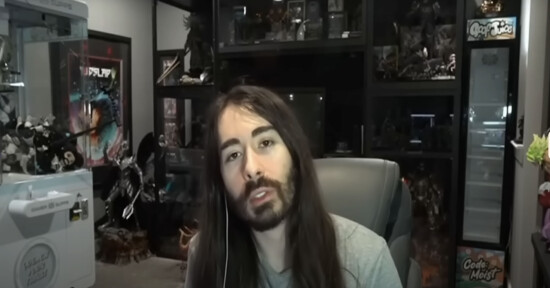 A person with long brown hair and a beard sits in a chair, speaking, with shelves filled with collectibles and action figures in the background.