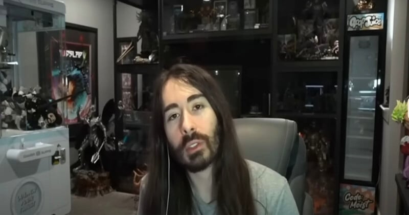 A person with long brown hair and a beard sits in a chair, speaking, with shelves filled with collectibles and action figures in the background.