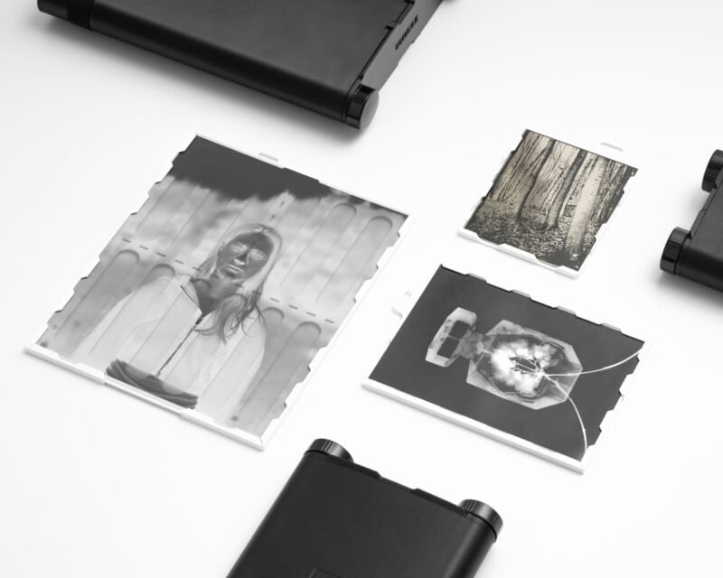 Three black-and-white photographic prints lie on a white surface, depicting a woman, trees, and a flower. Black rectangular cases are placed around the photos. The arrangement is minimal and orderly.
