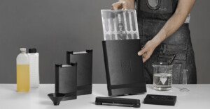 A person wearing a dark apron prepares film development chemicals and equipment on a white table, including black containers, bottles, a beaker, a measuring cylinder, a funnel, and a sheet of film being inserted into a black tank.