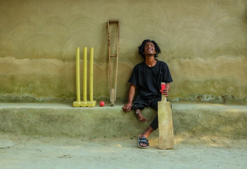A young man with a leg amputation sits on a low ledge, smiling joyfully, holding a cricket bat. Cricket stumps, a ball, and a wooden crutch rest beside him against a textured wall.