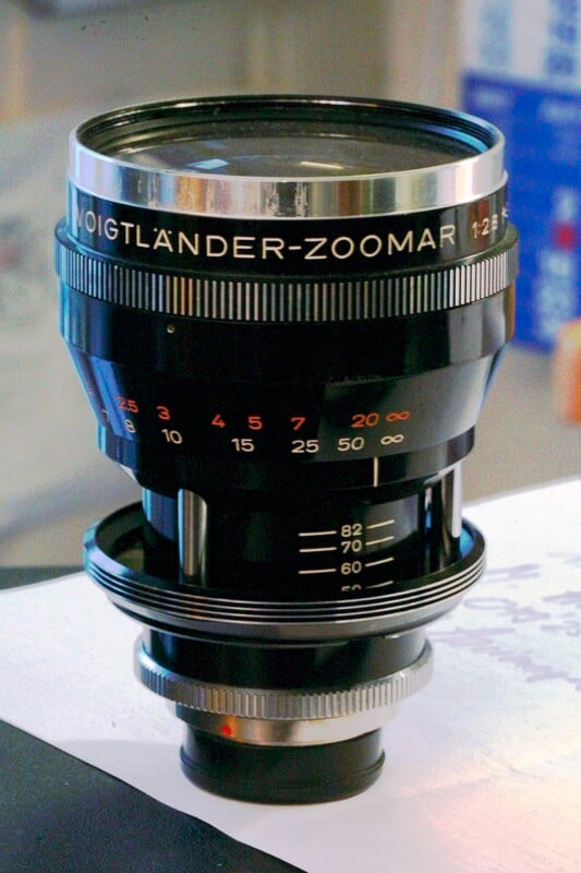 A close-up photo of a vintage Voigtländer-Zoomar camera lens with numbered focus markings and reflective metal details, placed on a white surface with a blurred background.