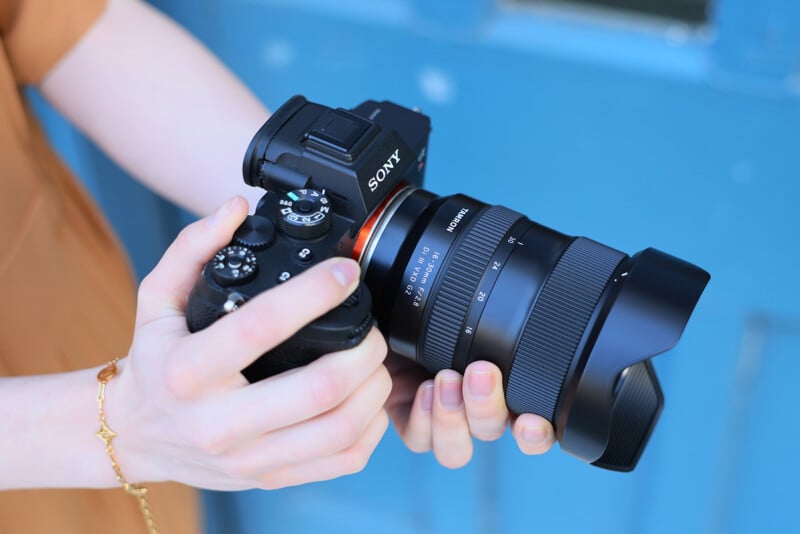 A person holding a black Sony digital camera with a large wide-angle lens, standing against a blurred blue background.