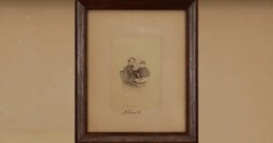 A sepia-toned photograph in a dark wooden frame shows two people sitting together. Below the photo on the beige mat, there is a handwritten signature that reads “A. Lincoln.”.