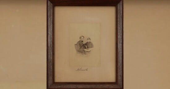 A sepia-toned photograph in a dark wooden frame shows two people sitting together. Below the photo on the beige mat, there is a handwritten signature that reads “A. Lincoln.”.