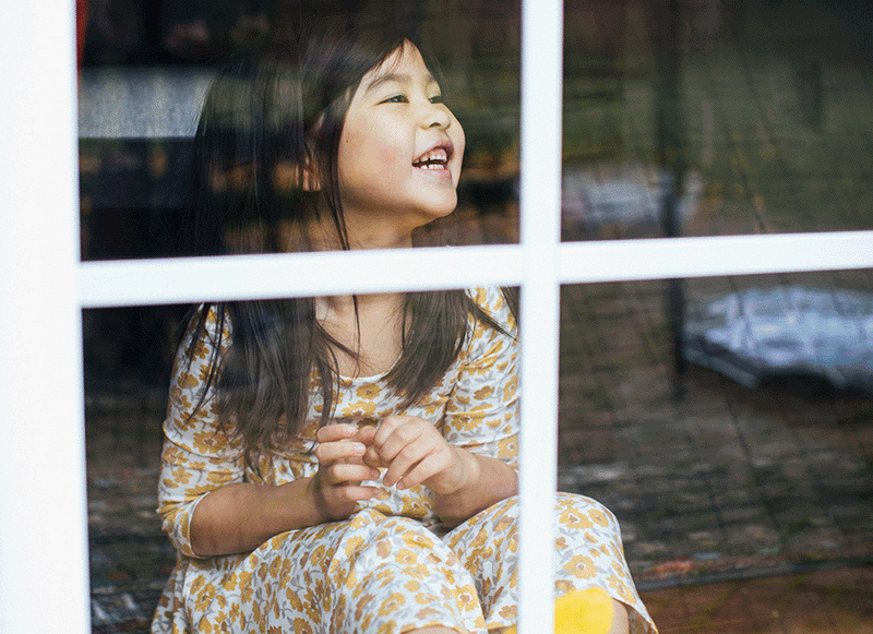 A young girl with long dark hair, wearing a yellow and white floral dress, sits smiling and looking up while behind a window with white grid panes. Reflections and outdoor scenery are visible on the glass.
