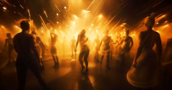 A group of people dance energetically under bright, warm yellow-orange lights, creating a blurred, dynamic effect that emphasizes movement and excitement.