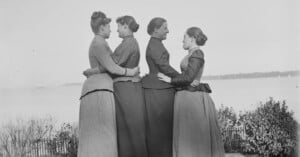 Four women in long skirts and jackets stand outdoors in a line, each facing sideways and embracing the woman in front of her. There is water and shrubbery in the background. The photo appears to be from the late 19th century.