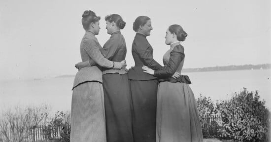 Four women in long skirts and jackets stand outdoors in a line, each facing sideways and embracing the woman in front of her. There is water and shrubbery in the background. The photo appears to be from the late 19th century.