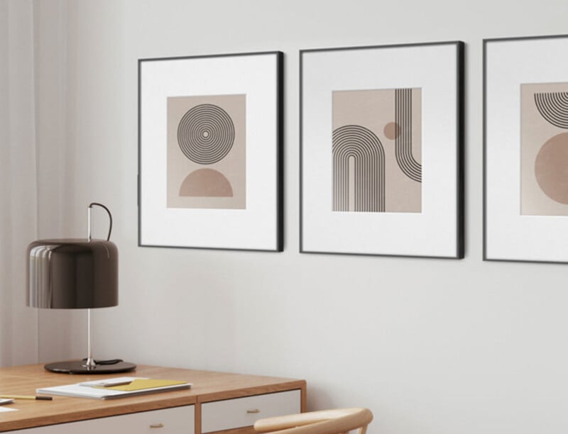 A modern desk with a dark lamp sits beneath three framed abstract art prints featuring black geometric shapes on beige backgrounds, hung on a white wall.
