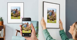 A woman hangs a framed photo of a man and child with a soccer ball on a wall. Next to it, someone uses a smartphone to scan the framed photo.